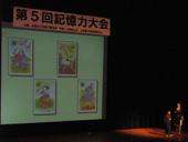 4枚のイラストカードが表示されたスクリーンと、舞台上で暗唱をしている発表者の写真