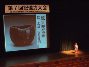 古い茶碗の写真と名前が映し出されたスクリーンと、舞台上に立つ女性の写真
