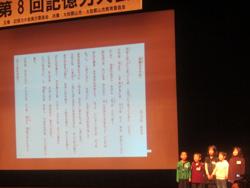 答えの文章が表示されたスクリーンを背に、舞台上で暗唱発表をする5人の子供達の写真