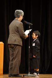 スーツ姿の女性が表彰状を読み上げる前で立っている、入賞者の男の子の写真