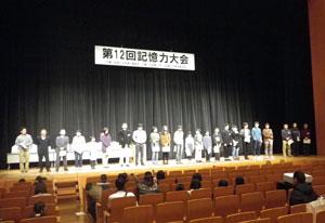 舞台上に横並びに立つたくさんの記憶力大会の参加者の写真