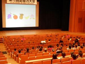スクリーンに映し出された果物のイラストを見ている客席の参加者達の写真