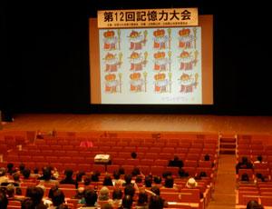 スクリーンに映し出された複数のイラストを見ている客席の参加者達の写真