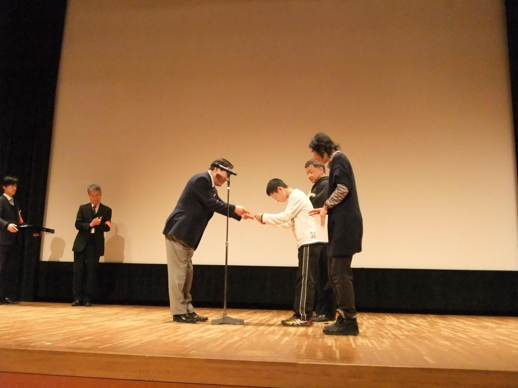 舞台上で男性から表彰状を受け取る、グループ3人組の写真