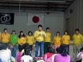 座っている子供達の前で、横に並んで立ち話をしている黄色い服をきた男女のグループの写真