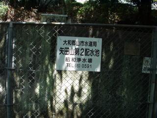 金網越しに見える石垣に貼られた「矢田山第2配水池」の看板の写真