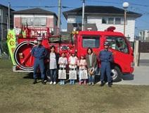 屋外で消防車の前に立って並ぶ消防団員と子供たちの集合写真