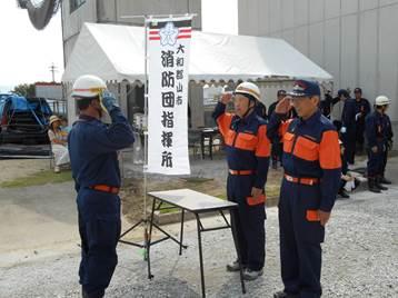 消防団指揮所と書かれた旗のそばで敬礼する消防団員三人の写真