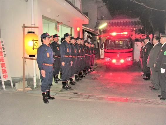 夜間に消防署前で整列する消防団員と消防車の写真