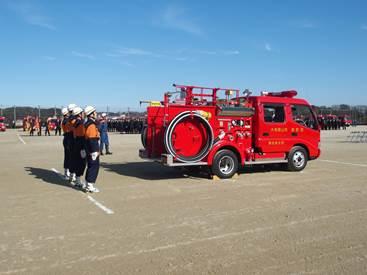 屋外に停車する消防車とその後ろで整列している消防団員の写真